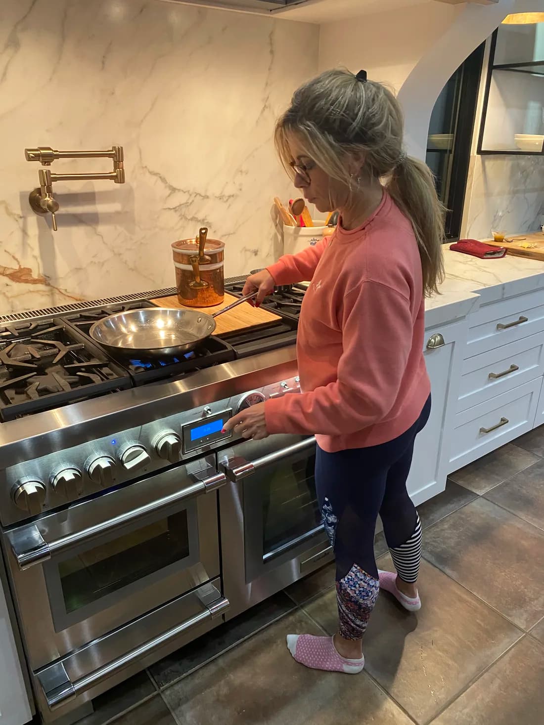 Andrea Williams at her home range in a coral sweatshirt, dialing in a pan over an open flame — grown-up cooking, seasoned like she means it.